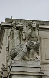 Stone statue of two figures: a man standing on the left with a heavy load on his shoulders a woman standing on the right in a helmet holding a ship's mast