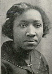 A young Black woman with her hair in a sidepart, wearing a dark collared blouse with a bar pin closure and contrasting stitching