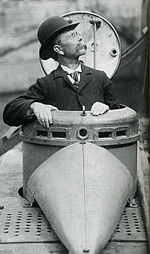 photographie en noir et blanc d'un homme à lunettes, moustachu et coiffé d'un chapeau melon, tourné vers la gauche dont le buste sort de l'écoutille d'un sous-marin