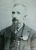 Head and shoulders of a white man with a long goatee, wearing a vest and suit coat with a flower pinned to the lapel.