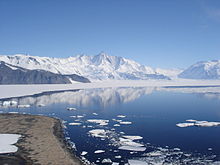 Montagne acérée enneigée devant une baie reflétant un ciel bleu.