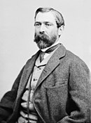 Black and white photo shows a bearded man seated. He wears civilian dress - a light colored vest and a darker coat.