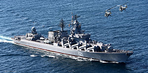 A large gray warship moves through open waters. Two helicopters fly overhead.