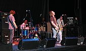 Five men on a stage; three in the forefront are holding guitars, while two in the background are behind a drum set and other equipment. Speakers, microphone stands and other equipment are also visible.