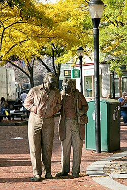 A sculpture resembling an elderly man and woman walking down the street, with trees and buildings in the background.