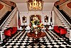 In a reception area, a large vase of flowers sits atop a wooden table in the center of a checkered black-and-white floor. Two staircases on either side flank the room.