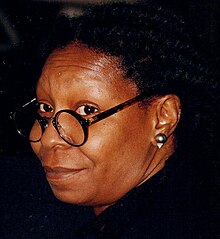 Middle-aged African-American woman with dreadlocks, looking away from the camera and smiling with her right cheek rested upon her folded hands.