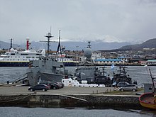 Imagem do porto da base naval mostrando 5 navios militares em primeiro plano e 2 navios maiores ao fundo, na distância além do porto se encontra uma cidade e um morro a direita.