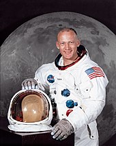 A portrait of a man wearing a space suit, with his helmet on the table in front of him. Behind him there is a large photograph of the lunar surface.