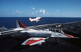 A TA-4 performs touch and go landings from Lexington, while another prepares to be catapulted from the flight deck in 1989