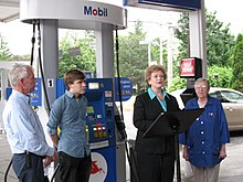 A white woman at a lectern on a Mobil gas station surrounded by another white woman and two white men.