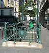 Vue en perspective se prolongeant vers le haut d'une bouche de métro, avec contraste entre le gris vert des ferronneries et le vert émeraude intense d'une poubelle placée dans le prolongement des escaliers.