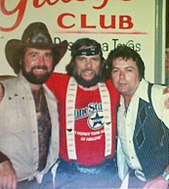 A group of three men with their arms round each other. The man on the left has a beard and is wearing a cowboy hat and a light-coloured vest. The one in the center has long hair and a beard and is wearing a bandana tied around his forehead, a red T-shirt and suspenders. The one on the right is wearing a white shirt and a dark vest.