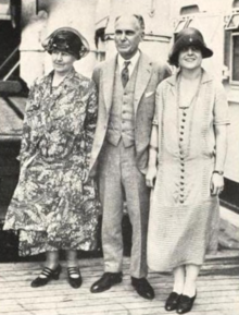 Three white people stand on a ship's deck for a photo: a older woman in a floral dress and dark hat, an older balding man in a suit, and a young woman in a cloche hat and dress with dark buttons down the front