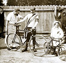 Photographie en noir et blanc d'un cycliste se tenant debout à côté de sa bicyclette, entouré d'un homme et d'un enfant.