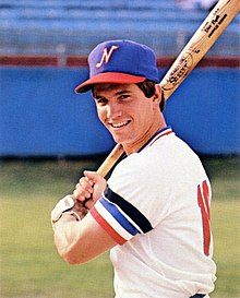 A man in a white baseball uniform with "Sounds" on the front in red and blue and a blue cap with a white "N" on the front posed holding his bat behind his head