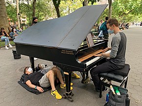 Huggins plays a grand piano on a large path in a park. Two people lie underneath it. On the piano's side is the phrase "THIS MACHINE KILLS FASCISTS".