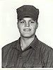 Head and shoulders of a smiling young white man wearing a cloth army cap and a plain buttoned-down shirt of the same color.