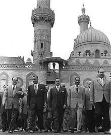 Several men walking forward, side by side. There are five men in the forefront, all wearing suits and ties. In the background is an ornate building with two minarets and a dome.