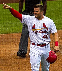 A man in a white baseball uniform with a red right sleeve holding a red helmet in his left hand points with his right hand.