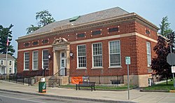 A one story brick building, seen from across the street and slightly to the right, with a peaked roof. It has decorative stone trim, with the words "U.S. Post Office" above the door.