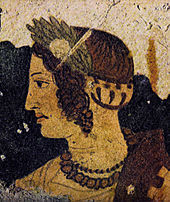 Détail d'une fresque de couleur marron, ocre, vert et noir représentant une tête de femme de profil aux cheveux bouclés, parée de deux colliers et portant une couronne de lauriers.