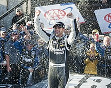 A man in his late thirties raising both his arms in the air to celebrate winning a race with a crowd of people behind him