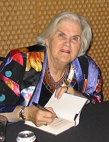 A woman with white hair wearing a light black and multicoloured jacket is seated at a table signing a book.