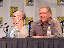 Two men seated in front of a table with microphones. The San Diego Comic Con logo can be seen in a panel behind them.