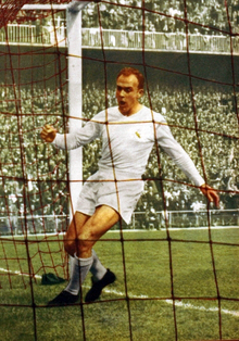 Photographie en couleur d'un footballeur habillé en blanc marchant à l'intérieur d'une cage de football, à l'arrière-plan des tribunes pleines de spectateurs.