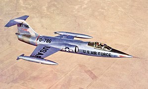 Jet fighter in metallic scheme with T-tail and short wings flying above desert and black constructions