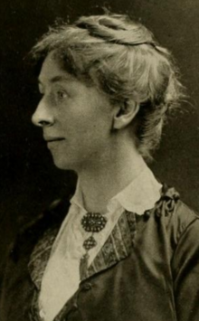 A white woman in 3/4 profile, wearing her long hair in an updo, a white blouse with a distinctive brooch pinned to the neck, and a darker jacket or overblouse