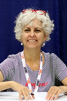 Kate DiCamillo at the 2018 US National Book Festival smiling at the camera holding a pen with red glasses resting on top of her head.
