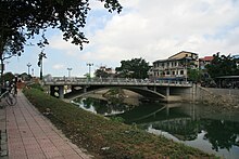 A small concrete footbridge, with an underarch, across a small body of water. It is for pedestrians and small lights are present. The sides of the water have a concrete embankment, and there is a footpath on the shore with a landscaped hedge.