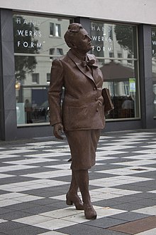 Photographie d'une statue en bronze marron dans une rue, représentant une femme marchant, un livre à la main.