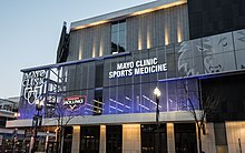 Four-story building clad in black, gray and beige-colored bricks, blue and white lighting, and the Mayo Clinic logo