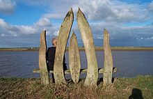 View from the rear of a seat made from 5 vertical rough-hewn planks of wood, with a person sitting on the seat and looking out over a wide river