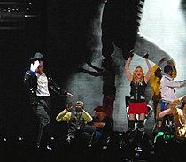 A group of males and a female performing on stage in front of a crowd of people. A male is shown blowing a kiss to the audience while wearing a white glove in his right hand and a white shirt with black pants and a jacket. An African American male wearing a white and dark gray shirt is shown in a crouched-down position. The female is waving her hands in the air while wearing black fingerless gloves with a black shirt and pink shorts. There are also two other African American males in the background making hand gestures. Behind the people on the stage, there is a screen that shows a black and white photo.