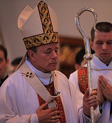 A man wearing a mitre, pallium, and chasuble holding a crozier in his left hand