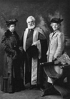 A bearded and elderly man dressed in a formal graduation robe posing with two female university representatives.