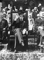 Masaryk, seated with his legs crossed and saluting the photographer
