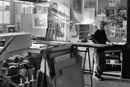 ANDRE COTTAVOZ chez Mourlot, debout, accoudé aux planches de travail dans la lumière de l'atelier.