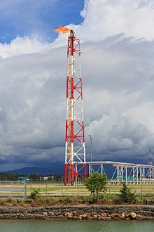 Des installations métalliques et des conduites blanches sont construites sur un terrain couvert d'herbe. En leur centre se trouve une torchère enflammée, dont les couleurs rouges et blanches contrastent avec le ciel aux nuages sombres. Au premier plan, des grillages barrent l'accès au site, et une montagne ainsi que des arbres sont visibles à l'arrière-plan.