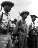 Three men, wearing uniforms and hats are standing and looking to the right. They are armed with grenades, guns, and have pouches. Other men can be seen in the background.