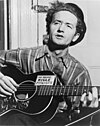 Black and white picture of a man in a plaid shirt and pin striped pants playing a guitar. A sticker with the phrase "This Machine Kills Fascists" appears on the guitar.