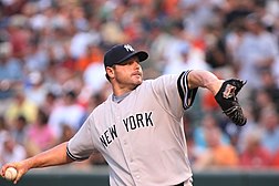 A man wearing a navy blue baseball cap with an interlocking "NY" and grey baseball jersey with "NEW YORK" on the chest prepares to deliver a pitch.