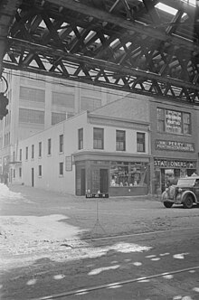 A black-and-white photograph taken across the street from two buildings, Costello's on the street corner and a stationary store on the right. The top foreground of the image shows light shining through the tracks of the Third Avenue El.