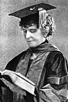 A white woman wearing academic cap and gown, gazing down at an open book in her hands