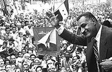 A man wearing a suit and tie with his upper body jutting out, waving his hand to crowds of people, many dressed in traditional clothing and holding posters of the man or three-striped, two-star flags