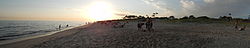 A panoramic photograph of a beach on a sunny day. There are people playing in the water, sitting in chairs, and walking in the sand along the water.